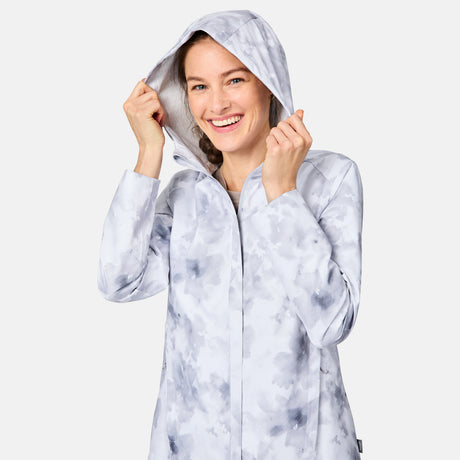 A smiling woman with braided hair wears the Free Country Women's X2O Packable Rain Jacket in a light gray and white pattern, holding both sides of the hood against a plain light background.