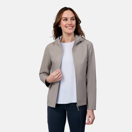 A woman with wavy brown hair smiles while wearing the Free Country Women's X2O Packable Rain Jacket in gray over a white shirt and dark pants, standing against a plain white background.