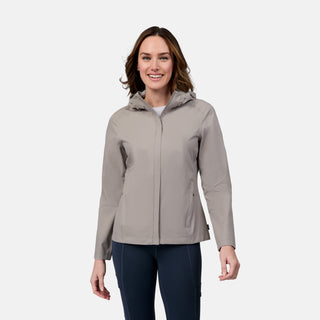 A woman smiles against a light gray background, wearing the Free Country Women's X2O Packable Rain Jacket—lightweight, breathable, waterproof, and UPF 50+—over a white top and dark pants.