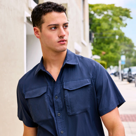 A young man with short dark hair wears a navy blue Clearance Men's Explorer Short Sleeve Shirt while standing on a sidewalk, looking to his left. Green trees and parked cars are visible in the background.