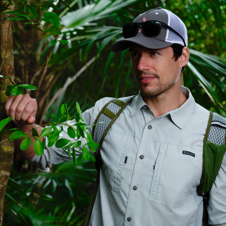 A man in a light gray Free Country Men's Acadia Long Sleeve Shirt, gray cap, sunglasses, and backpack stands in a lush green forest, holding a small tree branch while gazing into the distance.