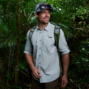 A man smiles in a lush, green forest wearing the Free Country Men's Acadia Long Sleeve Shirt in light gray, along with a baseball cap, sunglasses, and a backpack featuring green straps.