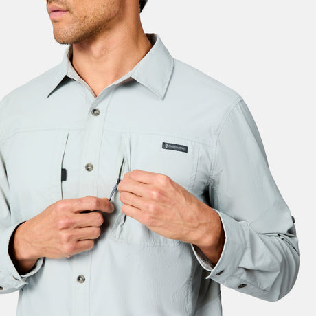 A man wears the Free Country Men's Acadia Long Sleeve Shirt in light gray, unzipping a chest pocket with his right hand. The shirt features a small rectangular label above the pocket and buttoned cuffs. His face is partially out of frame.