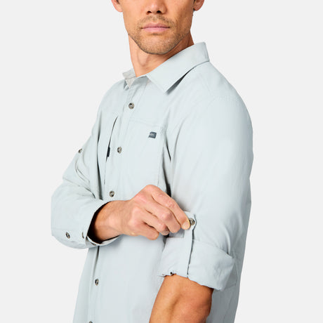 A man wearing a light gray Free Country Men's Acadia Long Sleeve Shirt rolls up his sleeve and secures it with a button tab. The background is plain white.
