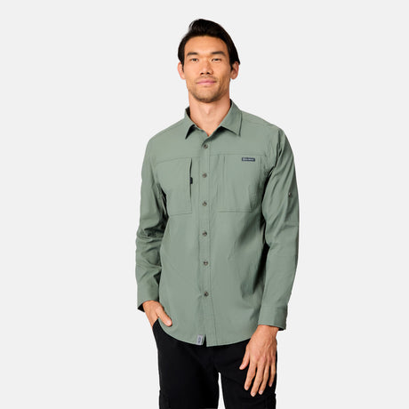 A man stands facing forward with one hand in his pocket, wearing a sage green Free Country Men's Acadia Long Sleeve Shirt with chest pockets and black pants, set against a plain light gray background.
