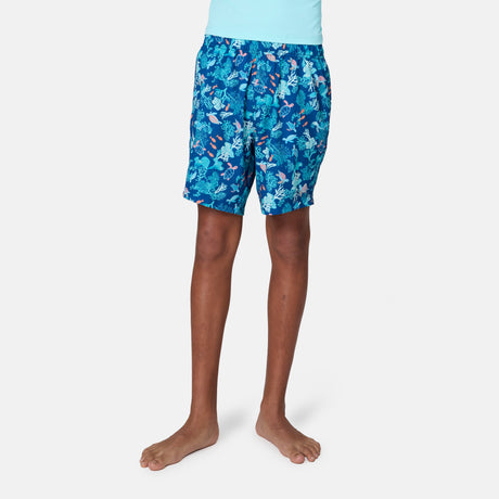 A boy stands barefoot in a light blue shirt and Free Country Boys' Reel Catch Swim Short, which features a colorful tropical fish and leaf pattern, against a plain white background.