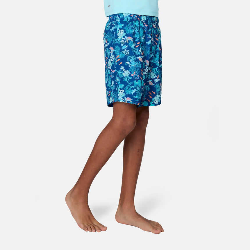 A boy stands barefoot in Free Country Boys' Reel Catch Swim Short, featuring a colorful ocean-themed pattern and quick-drying fabric, shown from mid-torso to feet against a plain white background.