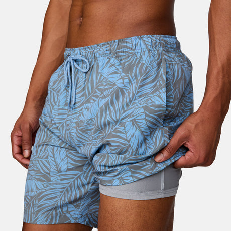 [Josiah is 6'3" wearing a size Medium.] Man wearing Free Country Men's Palm Heaven Swim Short in Bluestone Plam Heaven, size M, made from lightweight stretch recycled polyester performance fabric featuring a full elastic waistband with a drawcord, UPF and quick dry technology, and side seam pockets #color_bluestone-palm-heaven