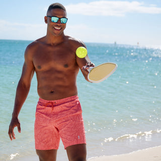 Wearing Free Country's Men's Paradise Palm Swim Short made from recycled polyester, a man enjoys a game of paddle ball on a sunny beach. He smiles behind his sunglasses with the ocean and blue sky in the background.