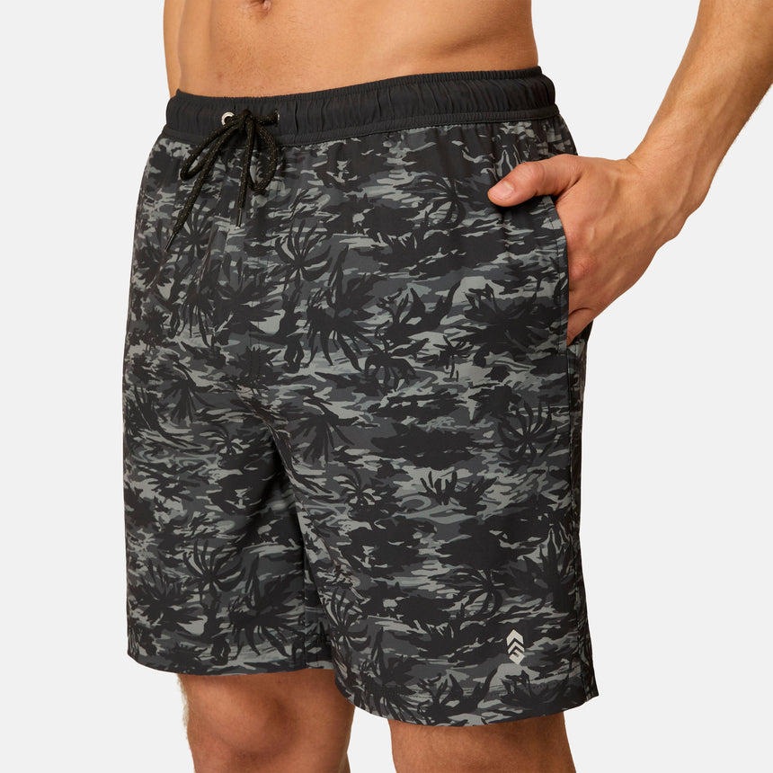A person is wearing the Free Country Men's Surf Camo Swim Short, featuring a black and gray leaf camo pattern and a drawstring waistband. Their right hand is in the pocket, and their torso is shirtless.