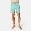 A person stands barefoot in Free Country Men's Surf Camo Swim Short—light teal with a white leaf pattern and drawstring waistband—against a plain white background.