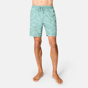 A person stands barefoot in Free Country Men's Surf Camo Swim Short—light teal with a white leaf pattern and drawstring waistband—against a plain white background.