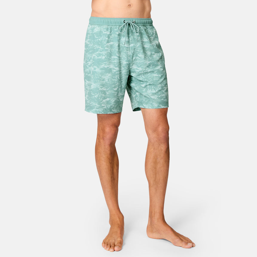 A person is wearing Free Country Men's Surf Camo Swim Short in light green with a subtle white tropical print and drawstring waistband, standing barefoot against a plain white background.