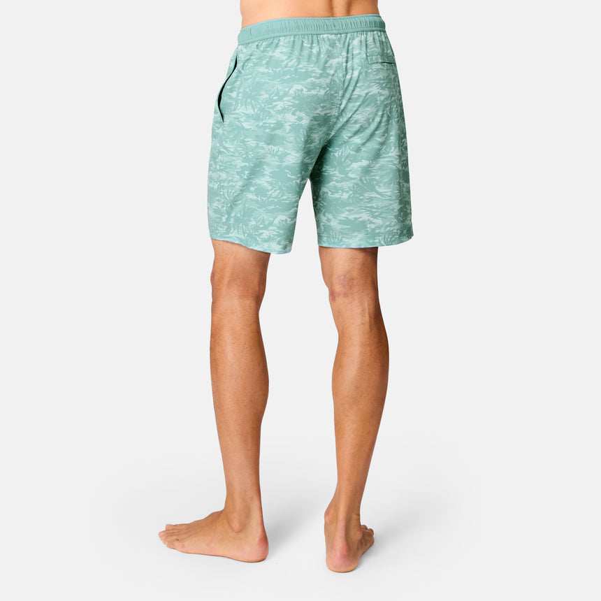 A person stands barefoot facing away, wearing Free Country Men's Surf Camo Swim Short featuring an elastic waistband and subtle camo pattern, set against a plain white background.