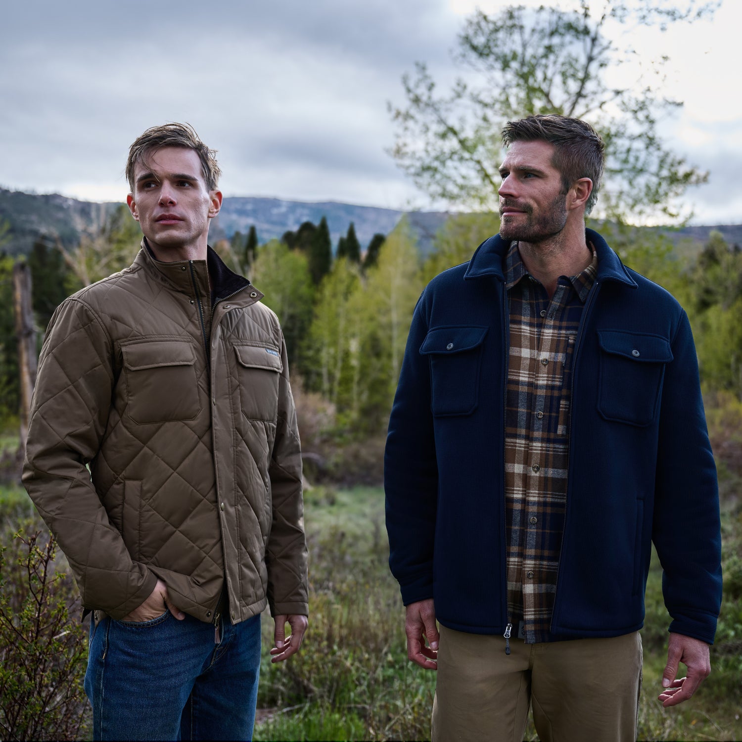 Two men stand outdoors in a forested area with trees and mountains in the background beneath a cloudy sky. One wears jeans and a brown quilted sherpa-lined jacket, while the other is dressed in khaki pants, a plaid shirt, and the Free Country Men’s Corded Fleece Shirt Jacket.