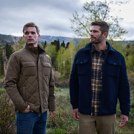 Two men stand outdoors in a forested area with trees and mountains in the background beneath a cloudy sky. One wears jeans and a brown quilted sherpa-lined jacket, while the other is dressed in khaki pants, a plaid shirt, and the Free Country Men’s Corded Fleece Shirt Jacket.