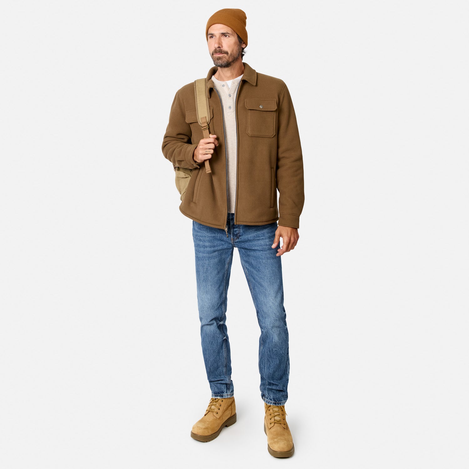 [Model is wearing size M.] A man wearing a brown overshirt, blue jeans, tan boots, and a tan beanie, carrying a beige backpack strap against a light gray studio background.