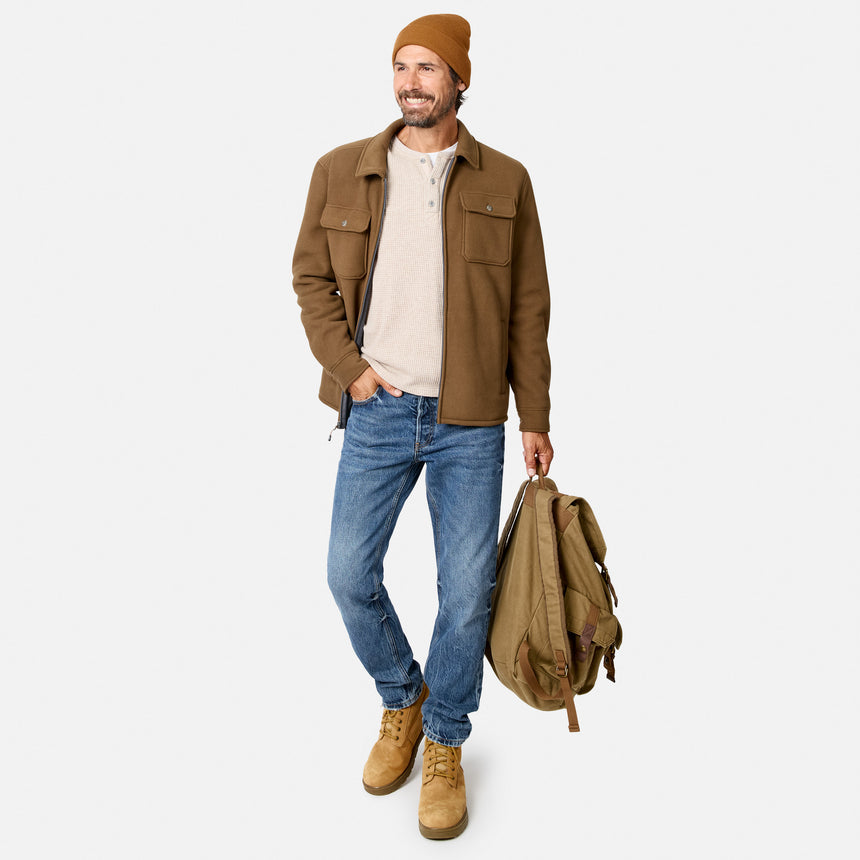 [Model is wearing size M.] A man wearing a brown jacket, cream henley shirt, blue jeans, and tan boots, holding a tan backpack against a light gray studio background.