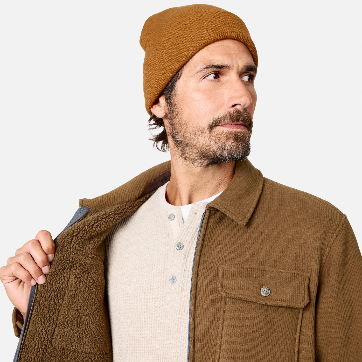 [Model is wearing size M.] A man wearing a tan ribbed knit beanie, a brown corduroy overshirt with a sherpa-lined collar, and a cream henley shirt, shown against a light gray studio background.