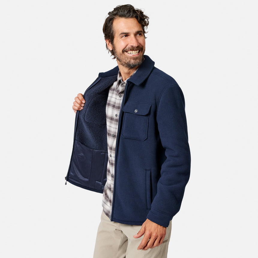 [Model is wearing size M.] A smiling man wearing a navy blue fleece jacket over a plaid shirt and khaki pants, holding the jacket open to show the fleece lining against a light gray background.