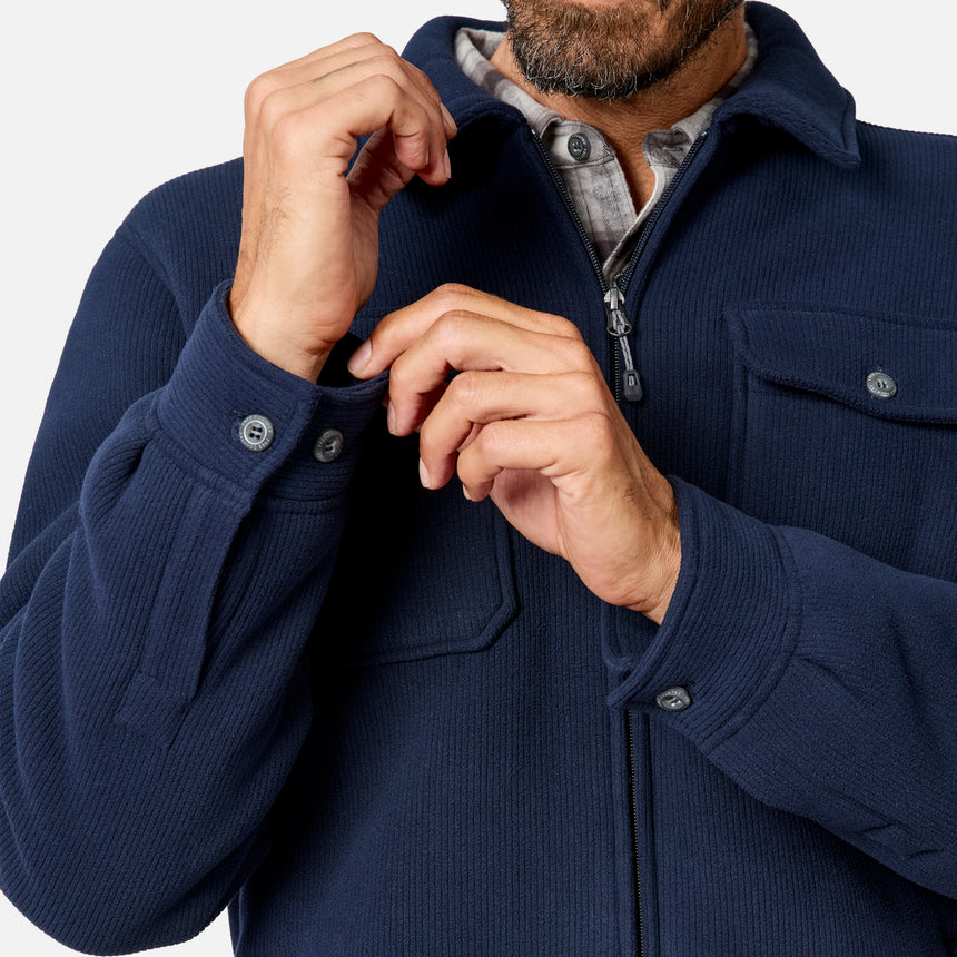 [Model is wearing size M.] A close-up of a man wearing a navy blue ribbed zip-up jacket over a plaid shirt, adjusting the collar and cuffs with his hands against a light gray background.