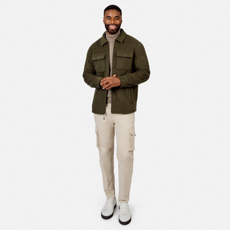 [Model is wearing size M.] A man wearing an olive green utility jacket over a beige turtleneck, paired with beige cargo pants and white sneakers, standing against a solid light gray background.