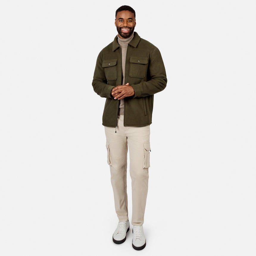 [Model is wearing size M.] A man wearing an olive green utility jacket over a beige turtleneck, paired with beige cargo pants and white sneakers, standing against a solid light gray background.