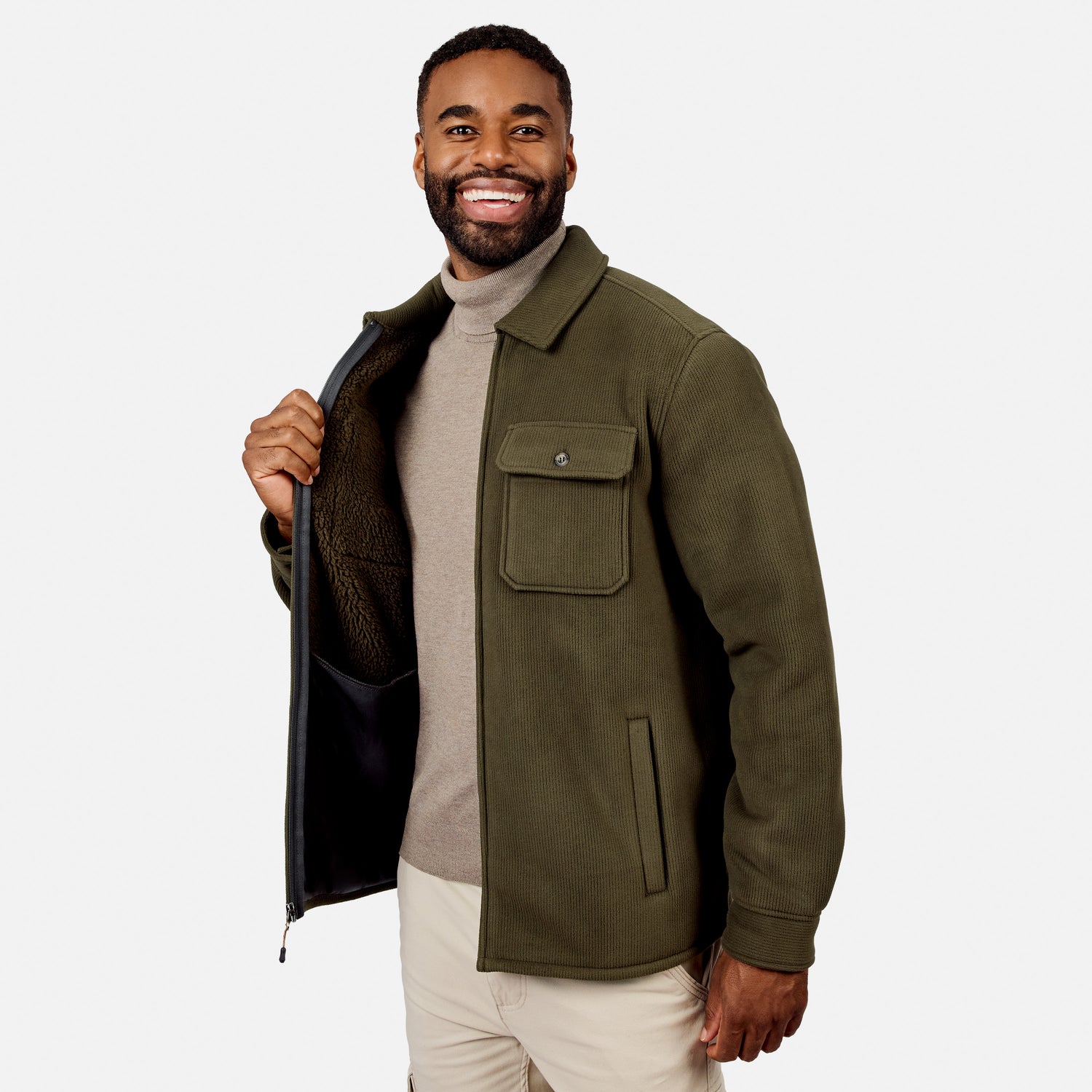 [Model is wearing size M.] A smiling man wearing an olive green fleece jacket over a beige turtleneck sweater and cream pants, holding the jacket open to show the dark sherpa lining, against a light gray studio background.