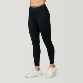 A person wears Free Country Women's Microtech® Heat Cocarber® Coffee Base Layer Bottoms (2-Pack) in black, paired with white athletic shoes, standing against a plain light background. The upper body is not visible.
