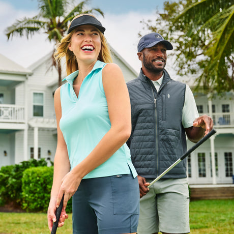 A woman in a visor and sleeveless top wears Free Country Women's Trail to Town Shorts while smiling with a man holding golf clubs outdoors on a manicured lawn, with houses and palm trees behind them.