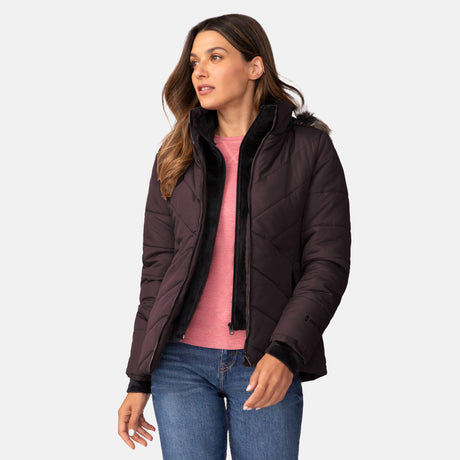 A woman in a Free Country Women's Brisk II Parka Jacket with a faux fur hood, water resistant and dark-colored, paired with a pink shirt and blue jeans, stands slightly turned left against a plain light background.