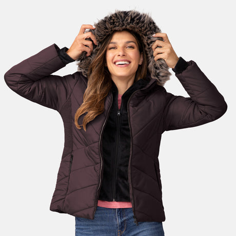 A woman with long brown hair smiles while holding up the faux fur-lined hood of the Free Country Women's Brisk II Parka Jacket, a dark, water-resistant winter coat. She pairs it casually with jeans.
