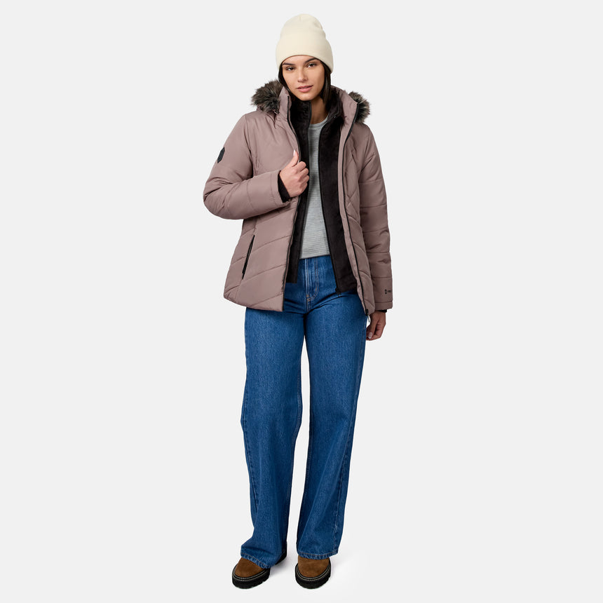 A person wearing a mauve Free Country Women’s Brisk II Parka Jacket with a faux-fur hood, a beige beanie, blue jeans, and brown boots.
