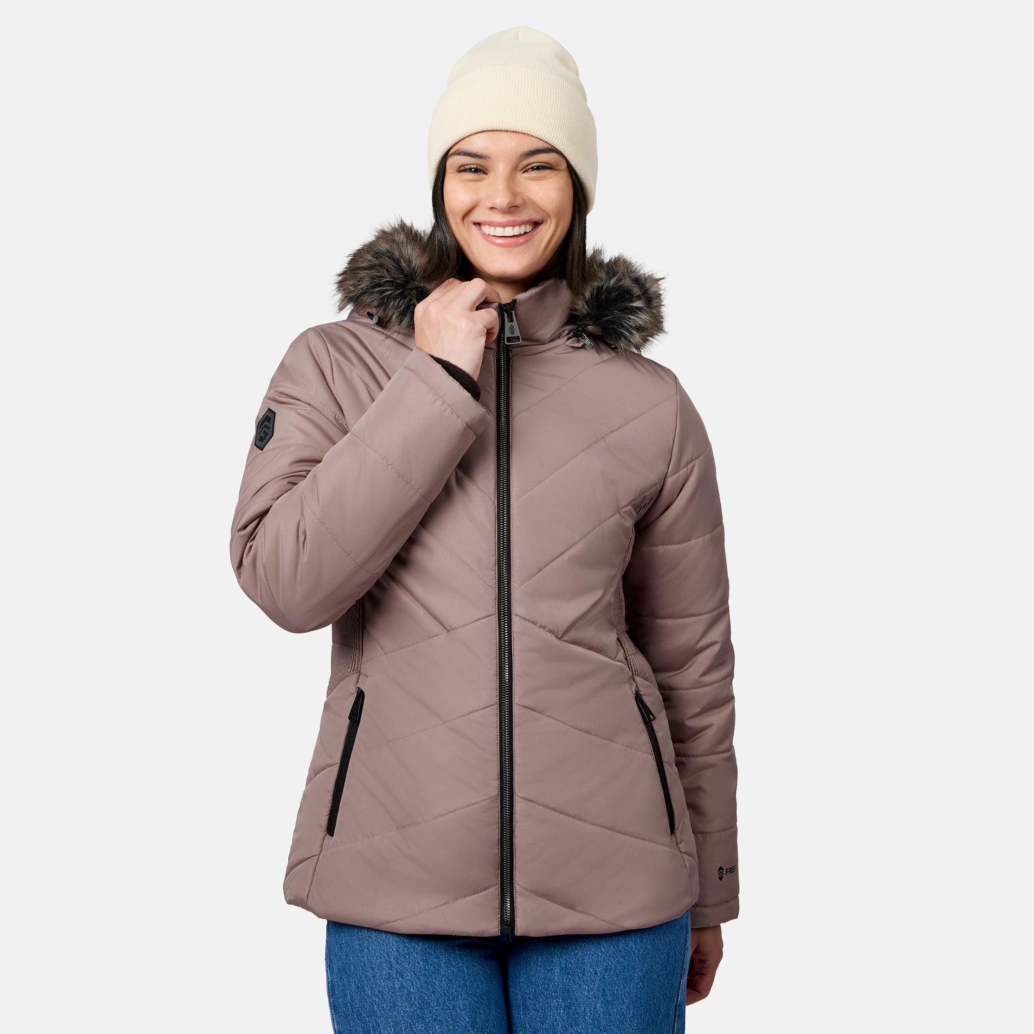 A woman wearing a tan beanie and a taupe quilted Free Country Women’s Brisk II Parka Jacket with a faux-fur-trimmed hood, zipped up to the chin.