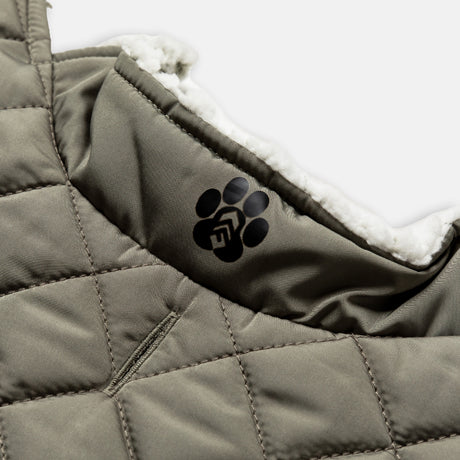 Close-up of the Clearance Stratus Lite Reversible Dog Jacket: a quilted olive-green coat lined with soft white fleece. A black paw print logo with a stylized F appears on the inside collar.