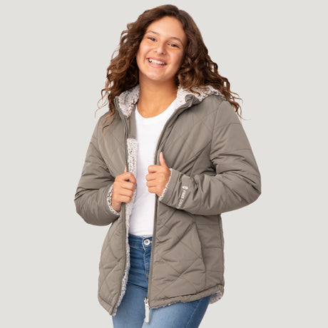[Model is wearing a size Medium.] Girl wearing the Free Country Girls' Stratus Lite Reversible Jacket in grey, featuring a diamond quilted pattern and a sherpa lining with a plain yolk, standing and smiling with hands in pockets against a plain backdrop.