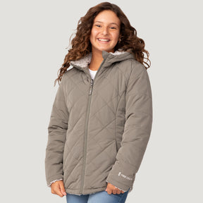 [Model is wearing a size Medium.] Girl wearing the Free Country Girls' Stratus Lite Reversible Jacket in grey, featuring a diamond quilted pattern and a sherpa lining, standing and smiling with hands in pockets against a plain backdrop.