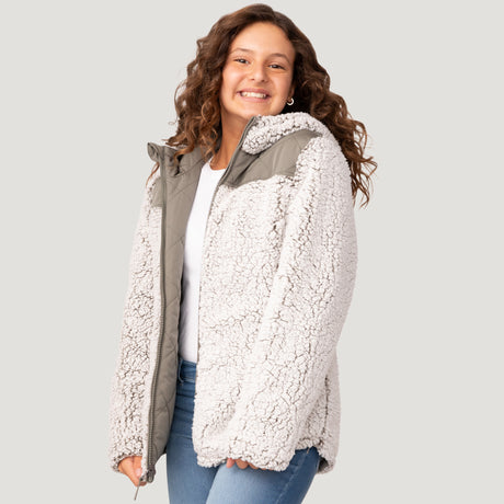 [Model is wearing a size Medium.] Girl wearing the Free Country Girls' Stratus Lite Reversible Jacket in grey, featuring a diamond quilted pattern and a sherpa lining with a plain yolk, standing and smiling with hands in pockets against a plain backdrop.