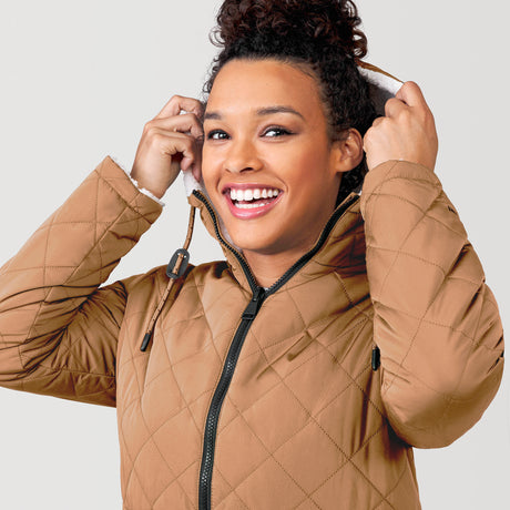 [Alexis is 5’6” wearing a size Small.] Woman wearing Free Country Women's Switch It Up Cloud Lite Reversible Jacket in tan with a full front zipper opening, attached hood with adjustable drawcord, diamond quilting, soft lining, and water resistant and wind resistant stretch performance fabric. light grey background. Perfect women’s  jacket for fall weather.