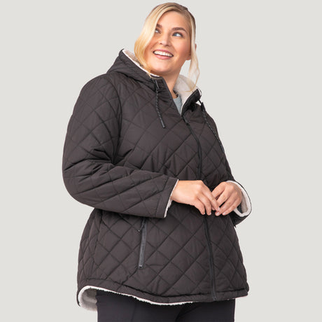 [Lola is 5'10.5" wearing a size 1X.] Woman wearing Free Country Women's Plus Size Switch It Up Cloud Lite Reversible Jacket in black with a full front zipper opening, attached hood with adjustable drawcord, diamond quilting, soft lining, and water resistant and wind resistant stretch performance fabric. light grey background. Perfect women’s  jacket for fall weather.