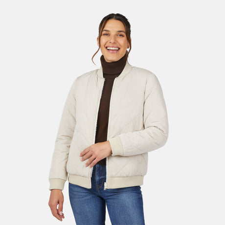 A smiling woman models the Clearance Women's Stratus Lite Reversible Quilted Bomber Jacket in cream over a dark brown turtleneck and blue jeans, standing against a plain white background.