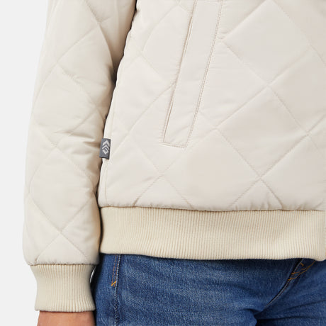 A close-up of a person wearing the Clearance Women's Stratus Lite Reversible Quilted Bomber Jacket in light beige with ribbed cuffs and hem, vertical pocket, small side tag, and blue denim jeans.