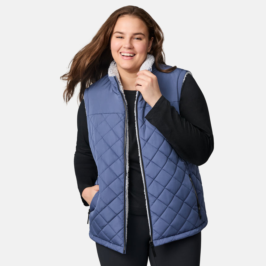 [Karissa is 5'9.5" wearing size 1X.] Women’s Free Country blue quilted vest with zip-front closure and zippered pockets—reverses to cozy sherpa for added warmth and texture. Worn over a black long-sleeve shirt and blue pants for a casual, transitional-weather look.