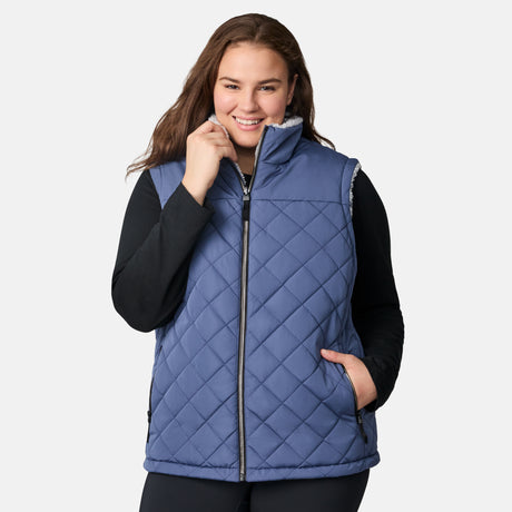 [Karissa is 5'9.5" wearing size 1X.] Women’s Free Country blue quilted vest with zip-front closure and zippered pockets—reverses to cozy sherpa for added warmth and texture. Worn over a black long-sleeve shirt and blue pants for a casual, transitional-weather look.