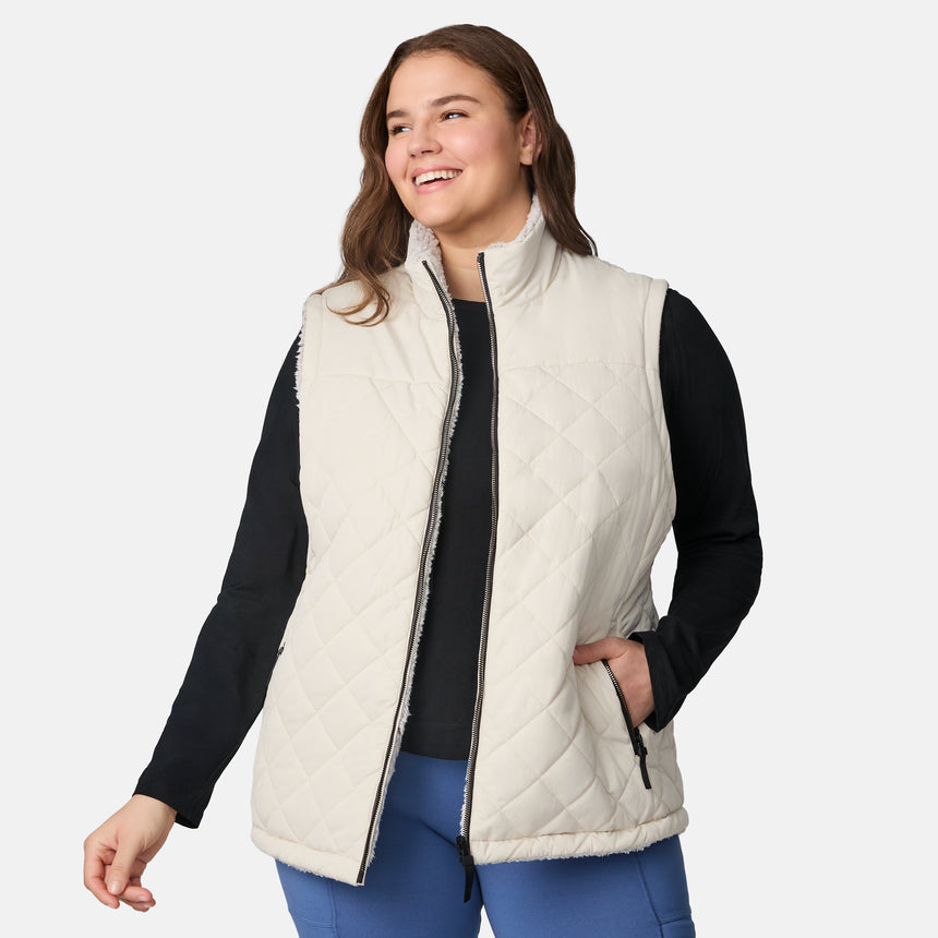 [Karissa is 5'9.5" wearing size 1X.] Women’s Free Country light beige quilted vest with zip-front closure and zippered pockets—reverses to cozy sherpa for added warmth and texture. Worn over a black long-sleeve shirt and blue pants for a casual, transitional-weather look.