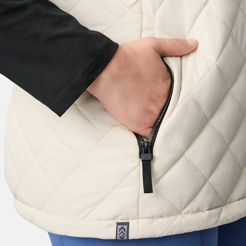 [Karissa is 5'9.5" wearing size 1X.] Women’s Free Country light beige quilted vest with zip-front closure and zippered pockets—reverses to cozy sherpa for added warmth and texture. Worn over a black long-sleeve shirt and blue pants for a casual, transitional-weather look.