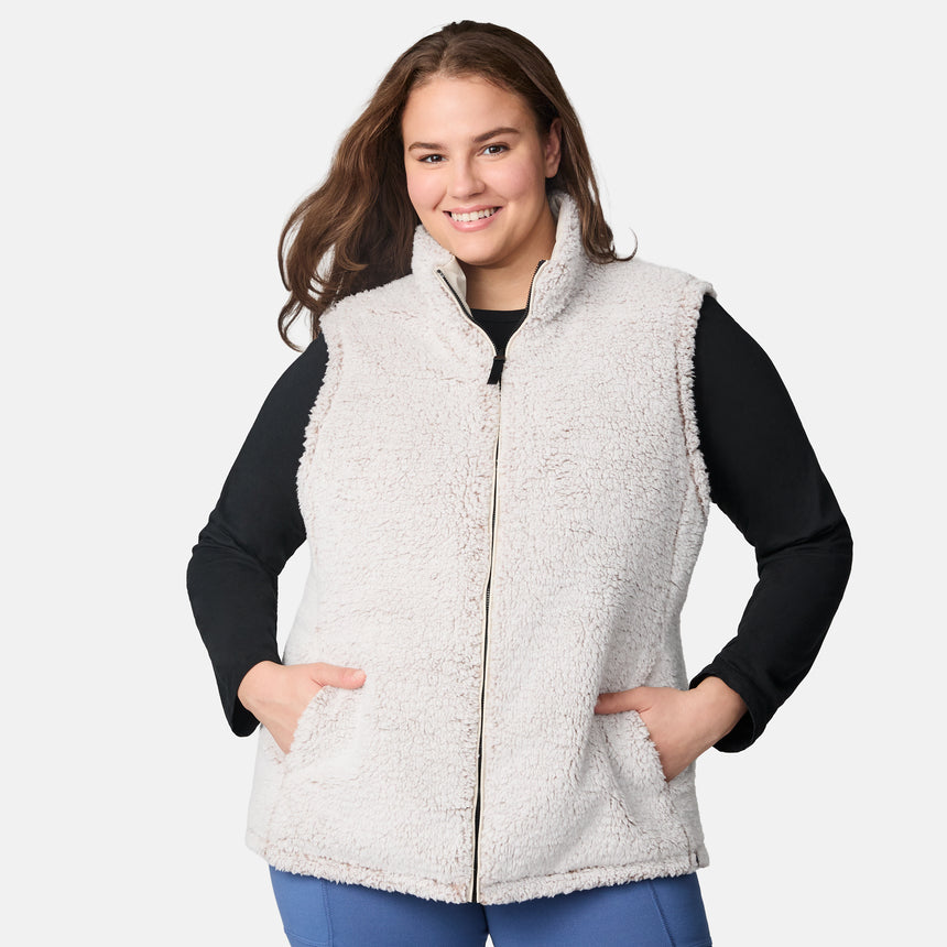 [Karissa is 5'9.5" wearing size 1X.] Women’s Free Country light beige quilted vest with zip-front closure and zippered pockets—reverses to cozy sherpa for added warmth and texture. Worn over a black long-sleeve shirt and blue pants for a casual, transitional-weather look.