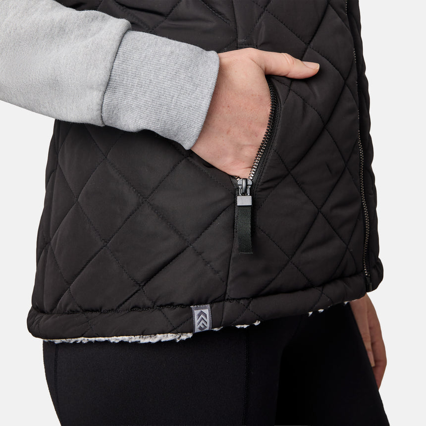[CJ is 5'10" wearing size S.] Women’s Free Country black quilted vest with zip-front closure and dual zippered pockets—reversible to cozy sherpa for added warmth and versatile styling. Layered over a light gray long-sleeve shirt for a casual, cold-weather look.