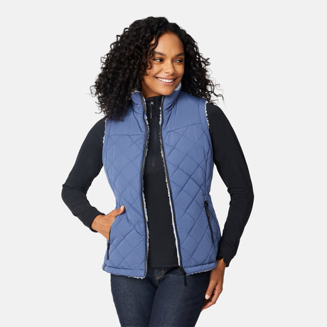 [Angelique is 5'10" wearing size S.] Women’s Free Country blue diamond-quilted vest with zip-front closure—reverses to cozy sherpa for added warmth and texture. Styled over a black long-sleeve shirt with one hand in pocket, set against a clean white background.