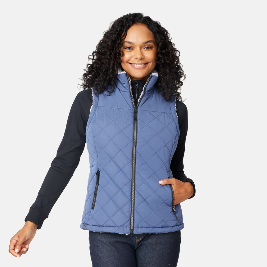 [Angelique is 5'10" wearing size S.] Women’s Free Country blue diamond-quilted vest with zip-front closure—reverses to cozy sherpa for added warmth and texture. Styled over a black long-sleeve shirt with one hand in pocket, set against a clean white background.